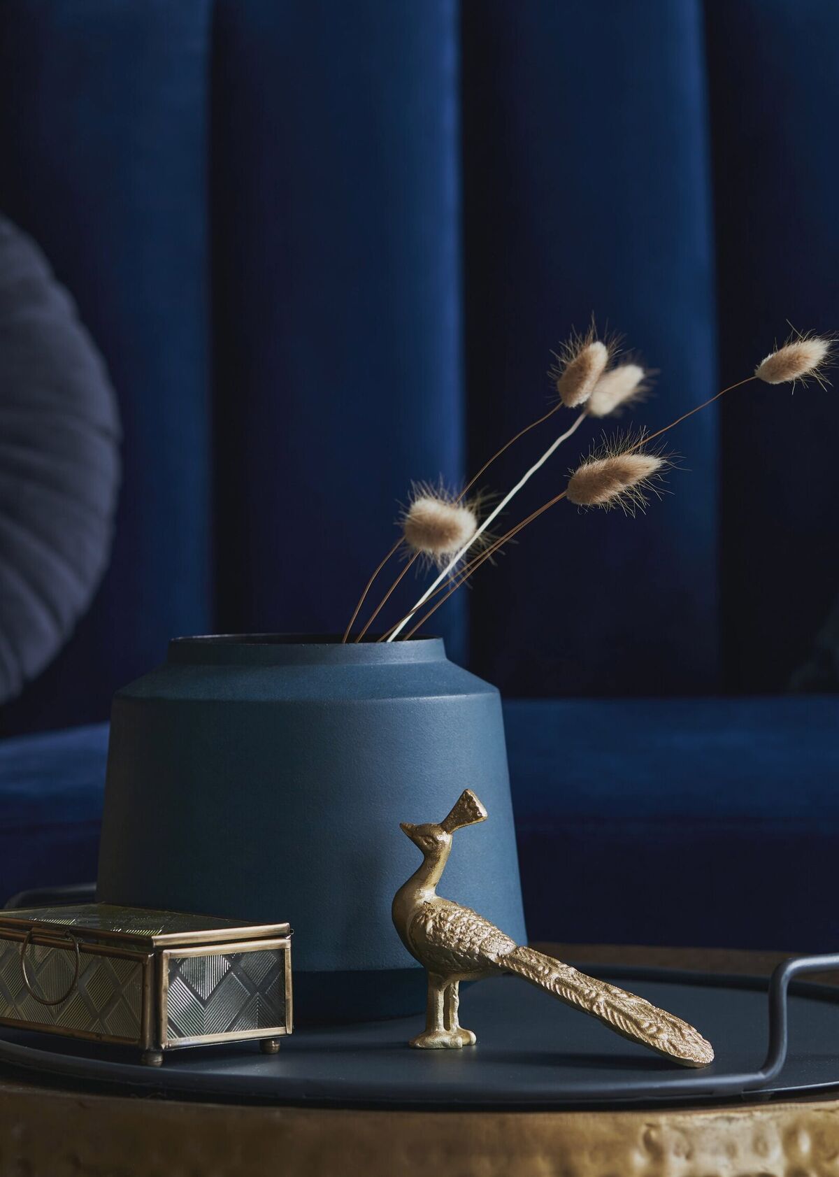 A brass peacock figure and a glazed box beside a matte vase of dried grasses, on a hammered brass table against blue velvet
