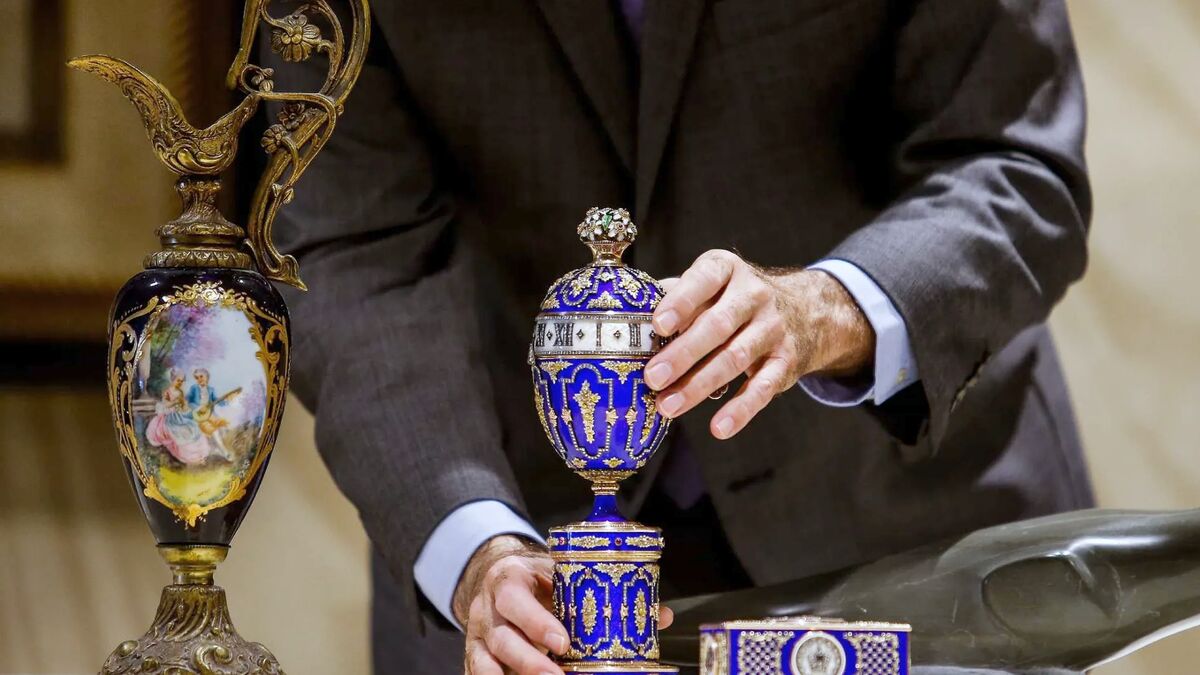 Hands in a suit steadying a cobalt and gilt enamelled Faberge-style egg beside a painted porcelain ewer