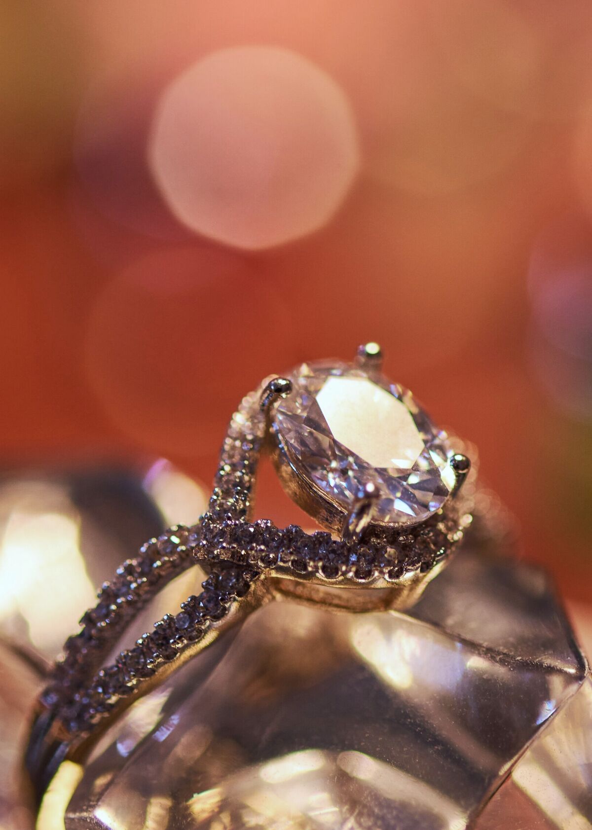 A diamond solitaire ring with a pave-set band, resting on cut glass against warm scattered highlights