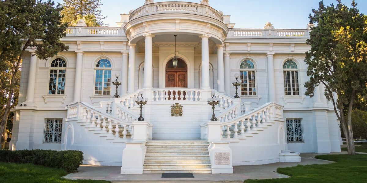 The columned portico and twin curving staircases of a white neoclassical mansion, seen from the garden path