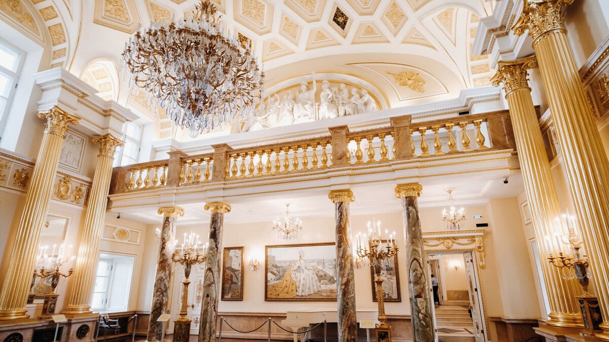 A palace ballroom with gilded Corinthian columns, a marble balustrade and a crystal chandelier beneath a coffered ceiling