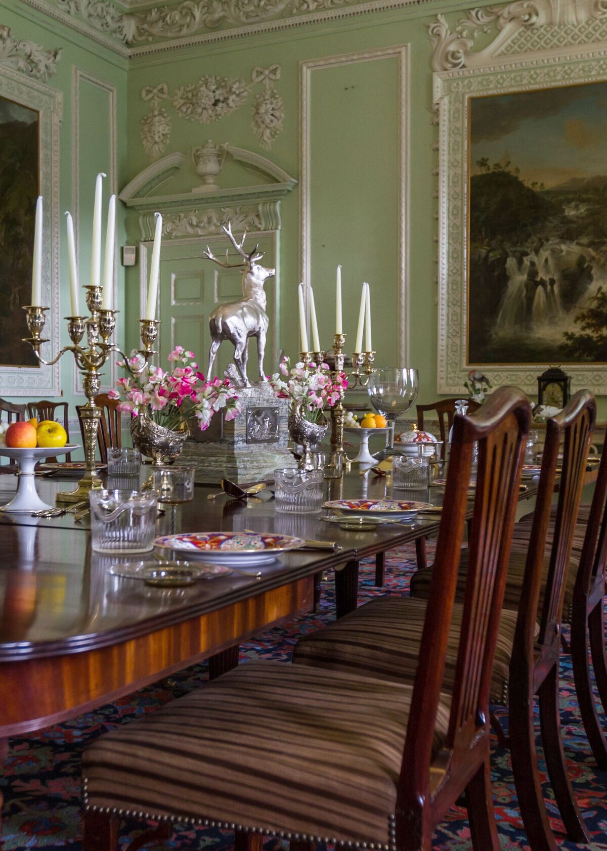 A long mahogany dining table laid with silver, crystal and candelabra in a green panelled room hung with oil paintings