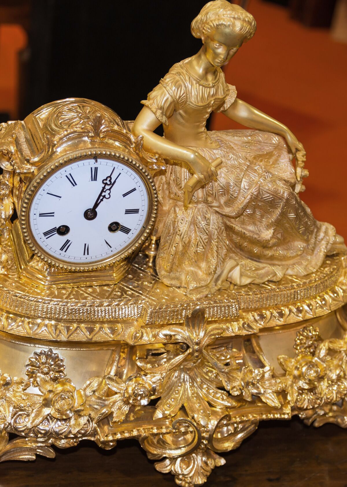 A gilt-bronze figural mantel clock, a seated woman resting against the enamel dial above a scrolled and foliate base