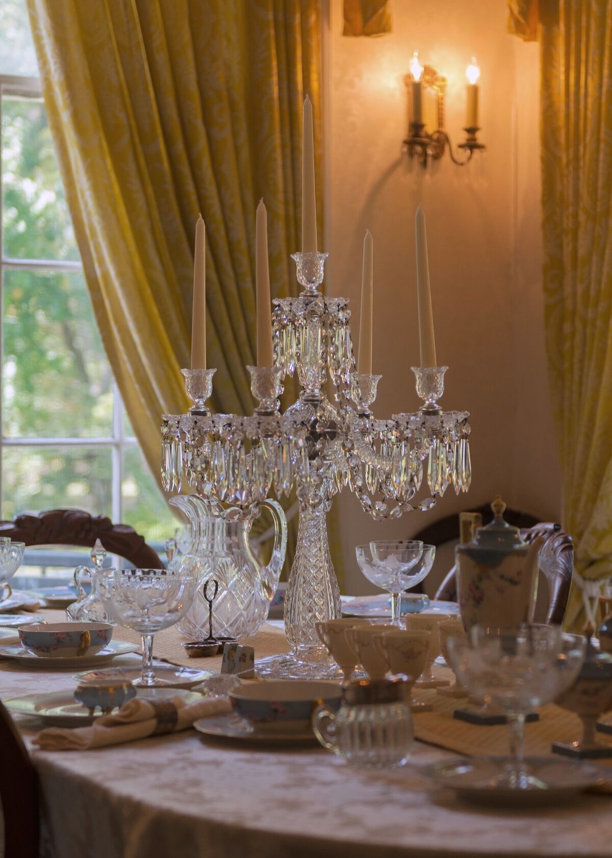 A dining table laid with cut crystal and painted porcelain around a crystal candelabrum, before tall windows hung with yellow silk