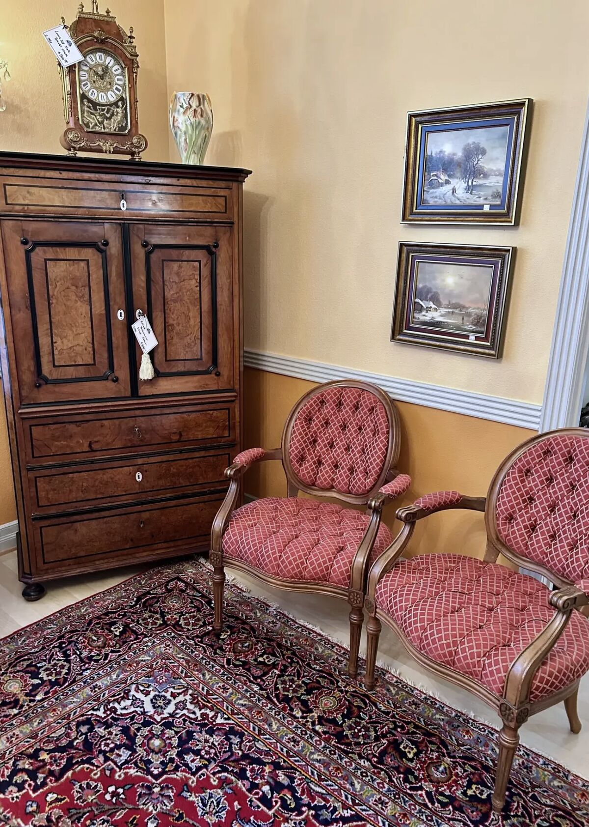 A burl-front chest and a pair of tufted salon chairs arranged on a Persian carpet, with a bracket clock and framed paintings above