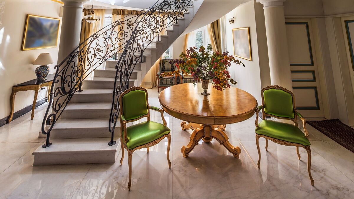 A grand two-storey foyer with a curving wrought-iron staircase, gilt side chairs and a round centre table beneath an ornamented ceiling
