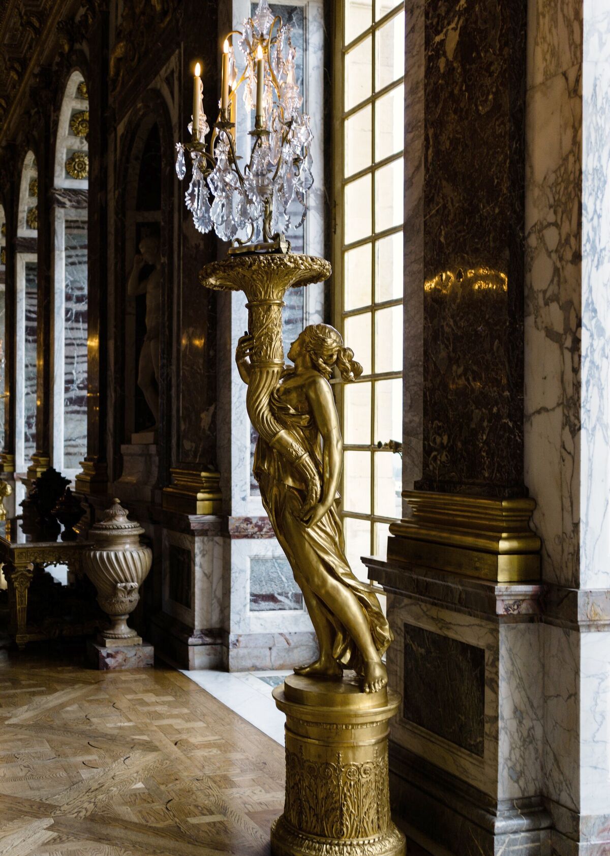 A gilded figural candelabrum standing against a marble pier beside a tall window in a palace gallery