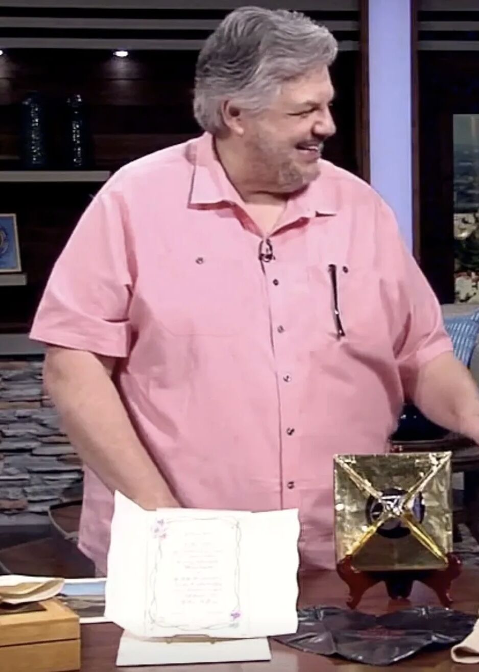 Gary Germer on the AM Northwest set, smiling beside a gold foil-wrapped pop-art piece and a framed certificate